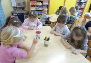 Pięcioro dzieci siedzi przy stoliku i koloruje sylwetę dziecka.